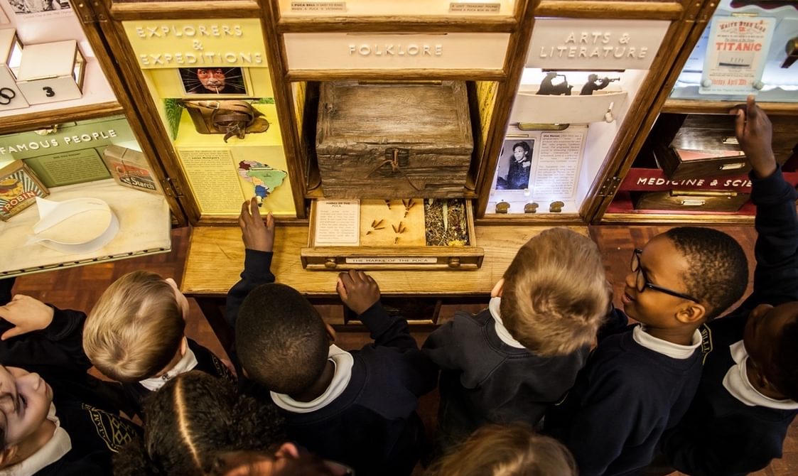 An overhead photo of a group of school children who are gathered around a cabinet of curiosities. The cabinet is split into different sections with titles like 'Folklore' and 'Arts and Literature'. In the centre is an old wooden box with a lock on it. One of the children is reaching out to touch the 'Explorers and Expeditions' section which includes a map.