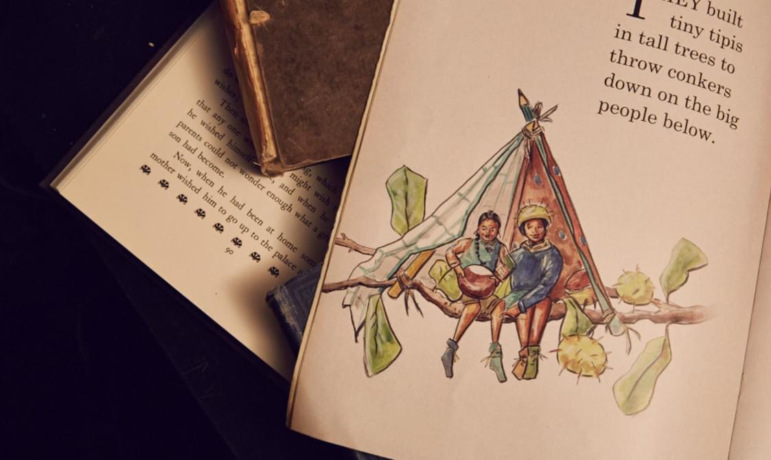 A photograph of an open storybook page. It has the text 'They built tiny tipis in tall trees to throw conkers down on the big people below'. You can see a drawing of two tiny people sat on a tree branch inside a tipi made with pencils and some cloth. Underneath is another open storybook and you can see text on the page.