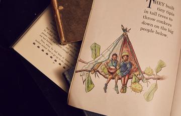 A photograph of an open storybook page. It has the text 'They built tiny tipis in tall trees to throw conkers down on the big people below'. You can see a drawing of two tiny people sat on a tree branch inside a tipi made with pencils and some cloth. Underneath is another open storybook and you can see text on the page.