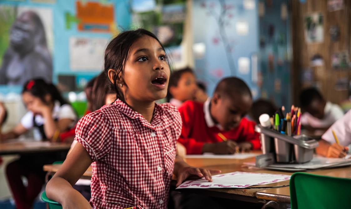 A girl is sat in a classroom, in the background you can see other children working. The girl in the foreground is wearing a red checked summer dress. She is holding a pencil in one hand, you can see a piece of paper on the desk. She is looking up in amazement with her mouth open. On the table you can see some pencils and glue in a grey plastic pot.
