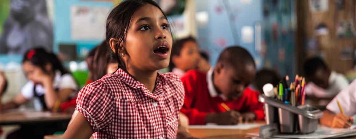 A girl is sat in a classroom, in the background you can see other children working. The girl in the foreground is wearing a red checked summer dress. She is holding a pencil in one hand, you can see a piece of paper on the desk. She is looking up in amazement with her mouth open. On the table you can see some pencils and glue in a grey plastic pot.