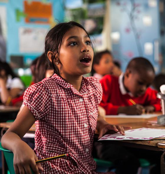 A girl is sat in a classroom, in the background you can see other children working. The girl in the foreground is wearing a red checked summer dress. She is holding a pencil in one hand, you can see a piece of paper on the desk. She is looking up in amazement with her mouth open. On the table you can see some pencils and glue in a grey plastic pot.