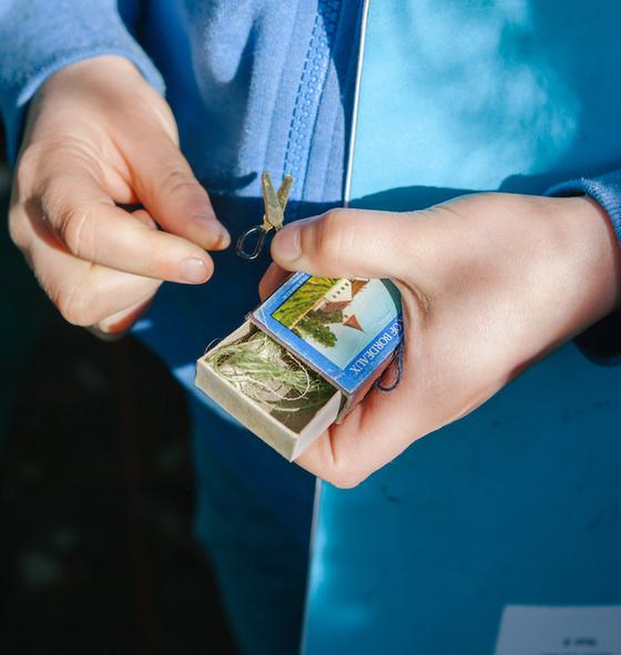 A close-up image of a child's hands. They are holding a matchbox which is half open, inside is some green string. The child is also holding a miniature set of scissors, it looks like they have been found inside the matchbox. The child is wearing a light blue zipped up hoodie and is holding a blue exercise book under their arm. The photo is taken outside, you can see some green grass in the background and sunlight is falling on their hands.