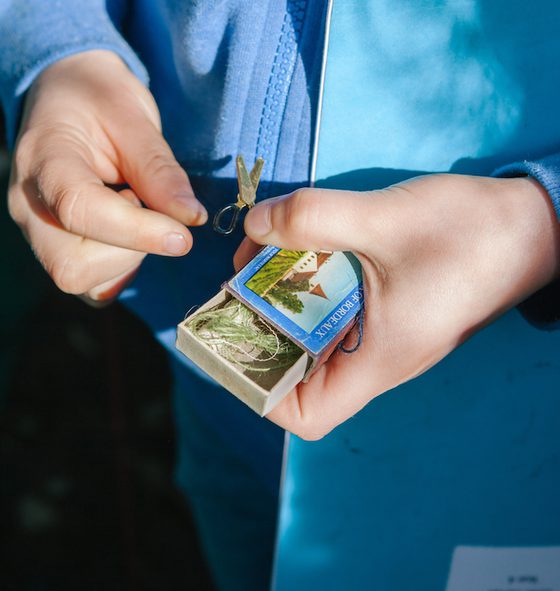 A close-up image of a child's hands. They are holding a mini box, inside is some green string. The child is also holding a miniature set of scissors, it looks like they have been found inside the matchbox. The child is wearing a light blue zipped up hoodie and is holding a blue exercise book under their arm. The photo is taken outside, you can see some green grass in the background and sunlight is falling on their hands.