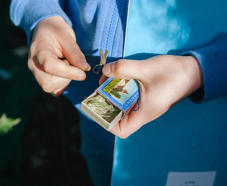 A close-up image of a child's hands. They are holding a matchbox which is half open, inside is some green string. The child is also holding a miniature set of scissors, it looks like they have been found inside the matchbox. The child is wearing a light blue zipped up hoodie and is holding a blue exercise book under their arm. The photo is taken outside, you can see some green grass in the background and sunlight is falling on their hands.