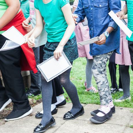 A full length image of school children stood together outside. You can't see their faces. They are all wearing different coloured clothes: a girl is wearing grey tights, black patent loafers, a blue skirt and green t-shirt. She is stood next to another girl wearing grey camouflage style leggings,  black shoes, white socks and a denim shirt. Each child is holding a sheet of white paper with a border. They look like they are rushing forwards.