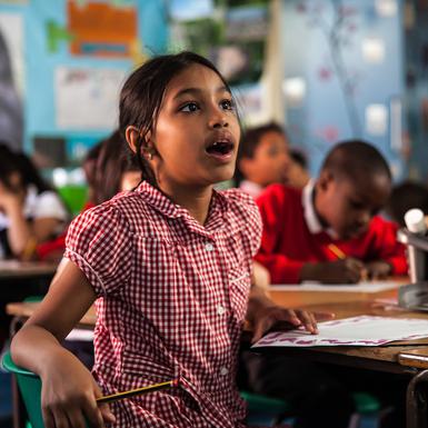 A girl is sat in a classroom, in the background you can see other children working. The girl in the foreground is wearing a red checked summer dress. She is holding a pencil in one hand, you can see a piece of paper on the desk. She is looking up in amazement with her mouth open. On the table you can see some pencils and glue in a grey plastic pot.