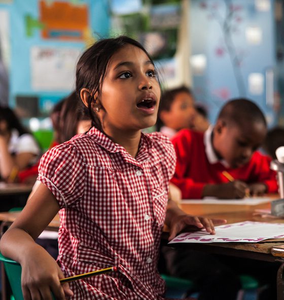A girl is sat in a classroom, in the background you can see other children working. The girl in the foreground is wearing a red checked summer dress. She is holding a pencil in one hand, you can see a piece of paper on the desk. She is looking up in amazement with her mouth open. On the table you can see some pencils and glue in a grey plastic pot.