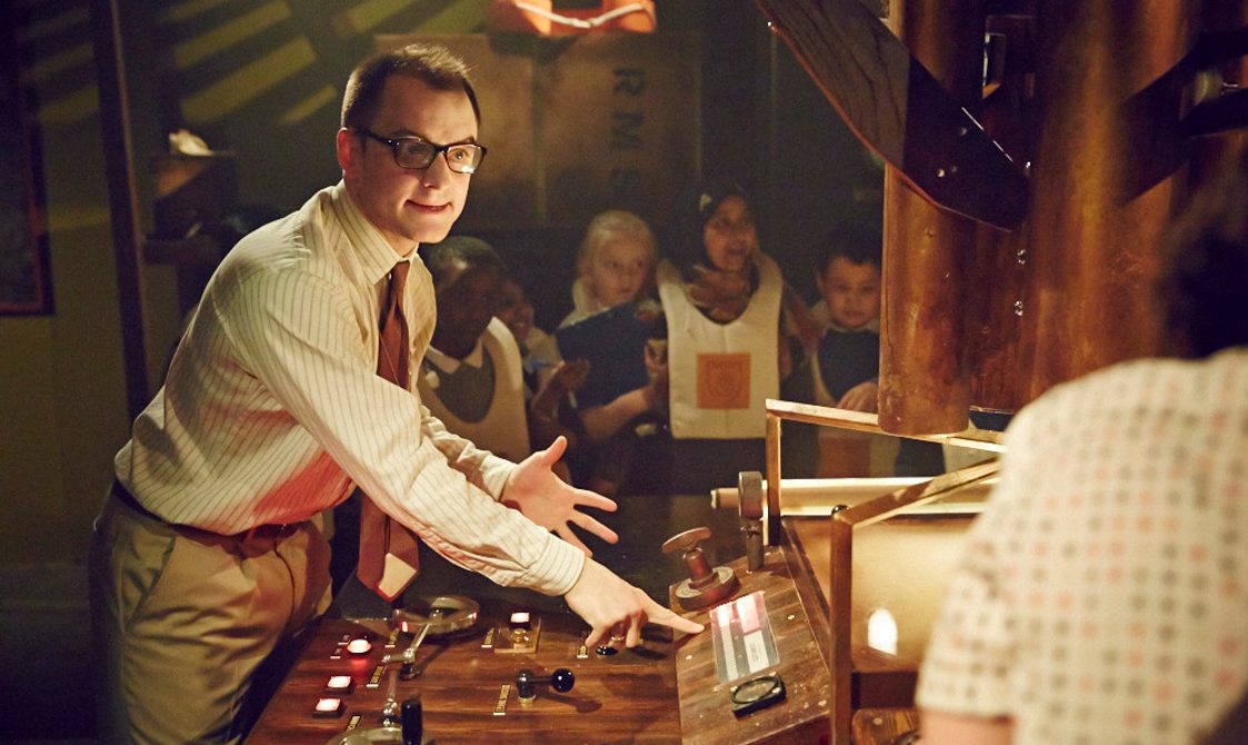 Image of a white man with short brown hair wearing glasses, a pinstripe shirt, red tie and light brown trousers. He is stood at a wooden control panel with brass buttons and red and white switches on it. In the background you can see children wearing old fashioned life jackets.