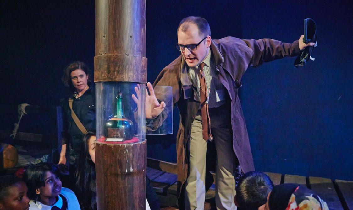 A man is wearing a dark brown trench coat, a faded red tie and a grey shirt with a badge on. His left hand is touching a glass cylinder case with an luminious turquoise jar inside. There are children looking up and around whilst on board a fantasy HMS Adventure.