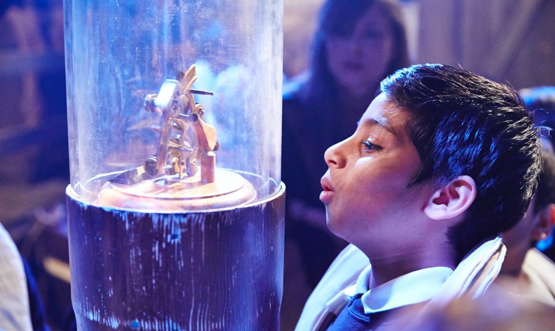 A young child is looking in awe and curiosity at a glass cylinder-shaped case with a wooden mechanical object inside.