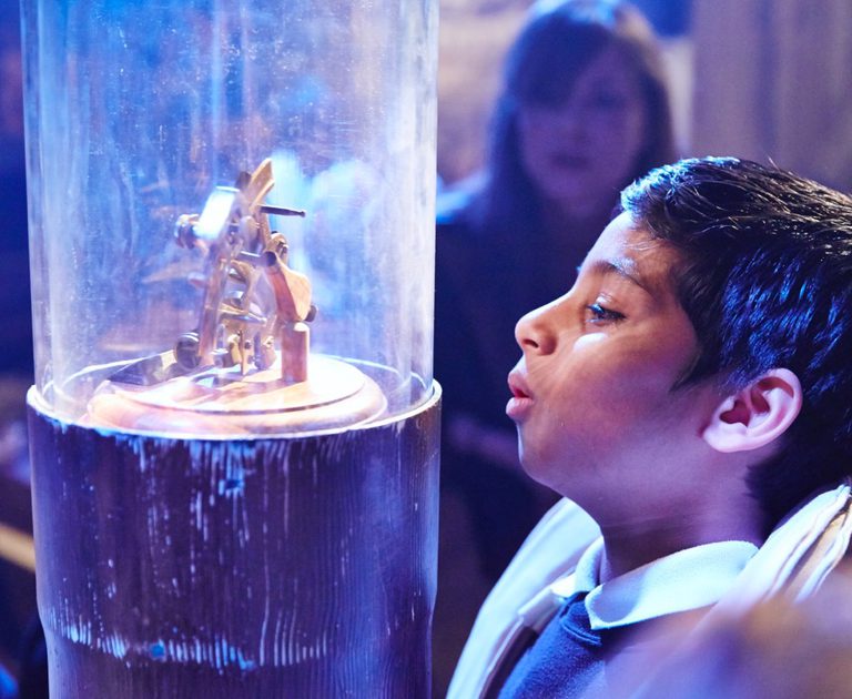 A young child is looking in awe and curiosity at a glass cylinder-shaped case with a wooden mechanical object inside.