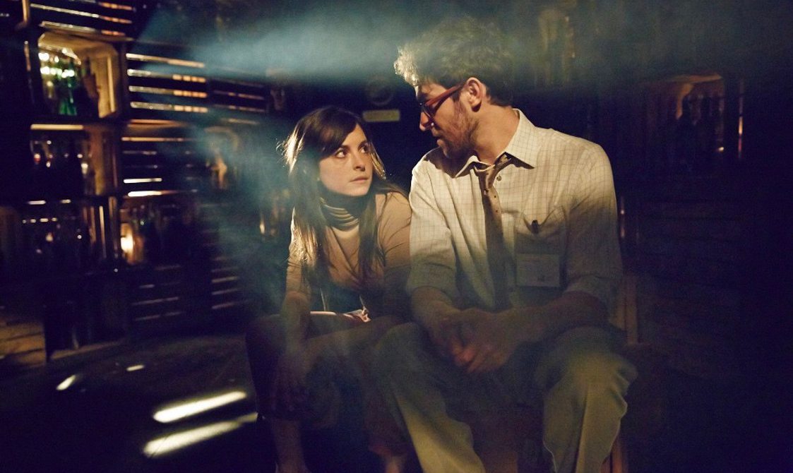 Two performers are sitting down looking at each other. They are in a darkly lit room with light shining out from the crated wall.