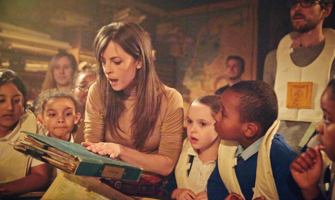 A woman is surrounded by a group of children who are all looking at the closed book she is holding with both of her hands. The children are in school uniform. The woman is wearing a brown jumper, and the book is a faded blue, and fairly large with old paper inside. The set design is inspired by a museum storehouse.