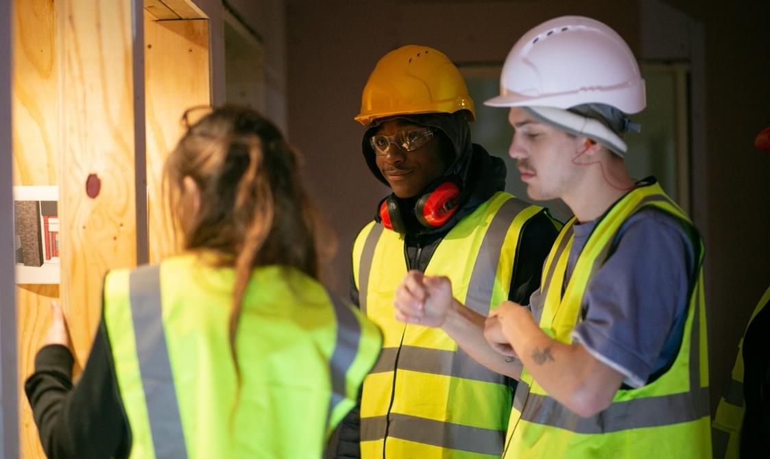 An image of two young men wearing a hi-vis and safety helmet. A woman wearing a hi-vis is facing away from the image and looking at a wooden structure.