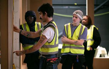 Four people are stood in a warehouse wearing hi-vis jackets. There is a man in the foreground side on motioning at a wooden wall. Two younger men are stood behind him looking on. Behind that is a woman watching what's happening.