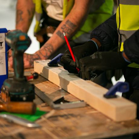 A close-up shot of two people doing carpentry at a wooden bench. A pair of hands in the foreground with gloves, using a red pencil to mark on a piece of wood. In the background are another pair of hands. Both people are wearing high-vis jackets. On the table is an electric drill, a chisel, a blue box and other tools that are out of focus.