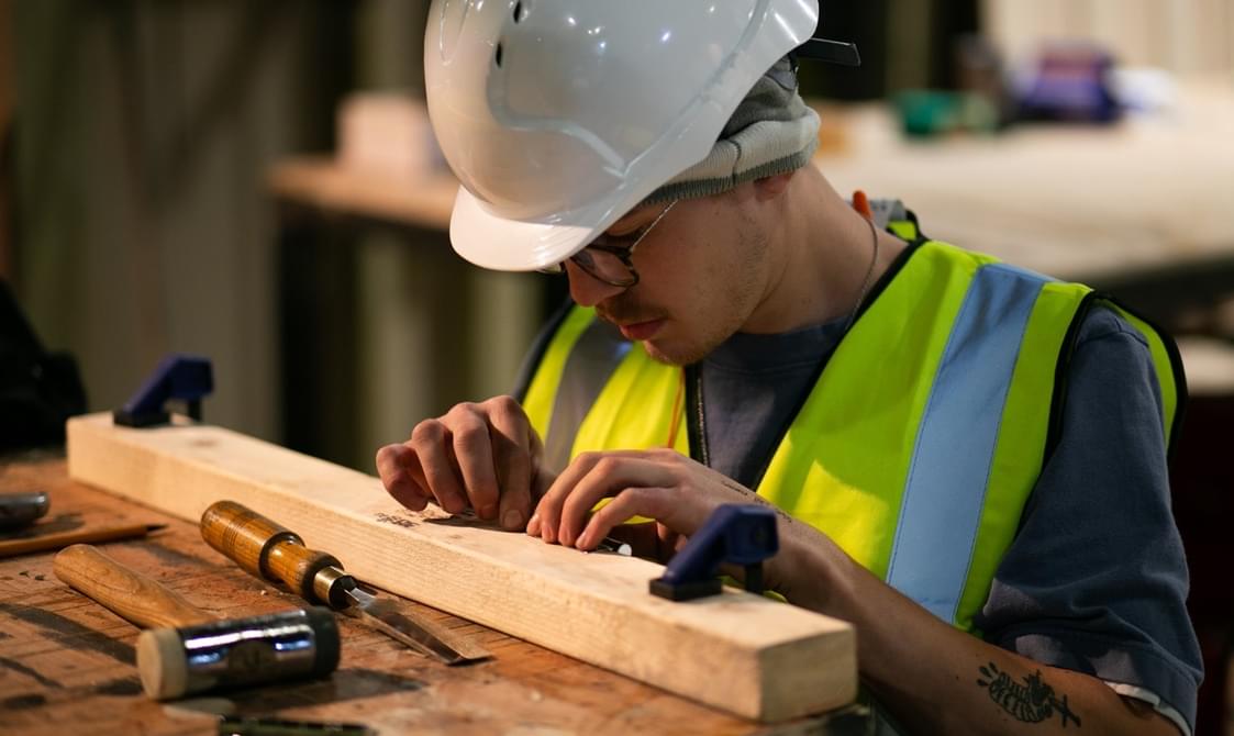 A young man wearing a high vis jacket and a white hard hat is kneeling at a wooden workbench. Both hands are on a piece of wood they are looking at. On the bench there is a chisel and a hammer.