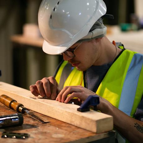 A young man wearing a high vis jacket and a white hard hat is kneeling at a wooden workbench. Both hands are on a piece of wood they are looking at. On the bench there is a chisel and a hammer.