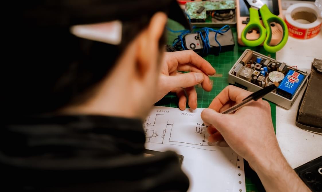 Overhead photo of a young man working at a desk. Their head and shoulders are out of focus but you can see they are wearing a black baseball cap and a black jumper. They are using a black biro to draw a a plan on a white piece of paper. Around them on the desk you can see a green handled pair of scissors, orange tape, a green cutting matt and the inside of an electrical item.