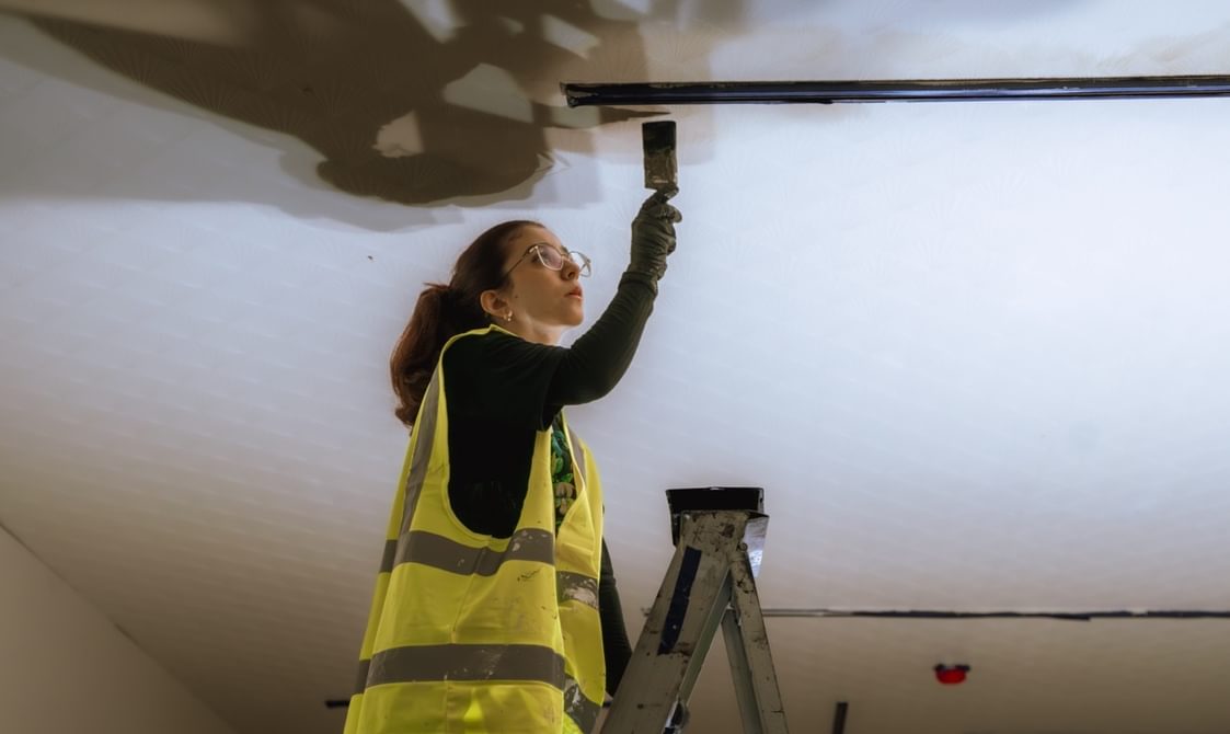 A woman wearing a high vis jacket and glasses is stood at the top of a ladder painting a ceiling.