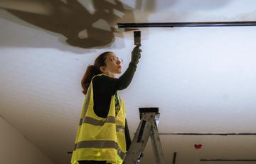 A woman wearing a high vis jacket and glasses is stood at the top of a ladder painting a ceiling.