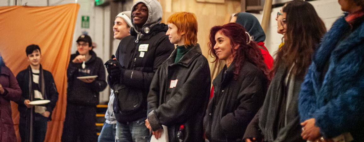 A group of young people stand in a line in a warehouse. They are smiling and all wearing coats.