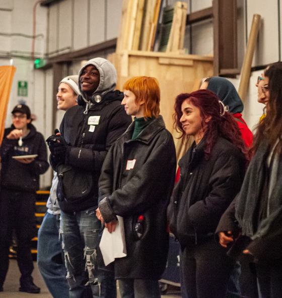 A group of young people stand in a line in a warehouse. They are smiling and all wearing coats.