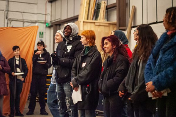 A group of young people stand in a line in a warehouse. They are smiling and all wearing coats.