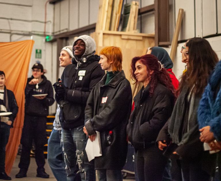 A group of young people stand in a line in a warehouse. They are smiling and all wearing coats.