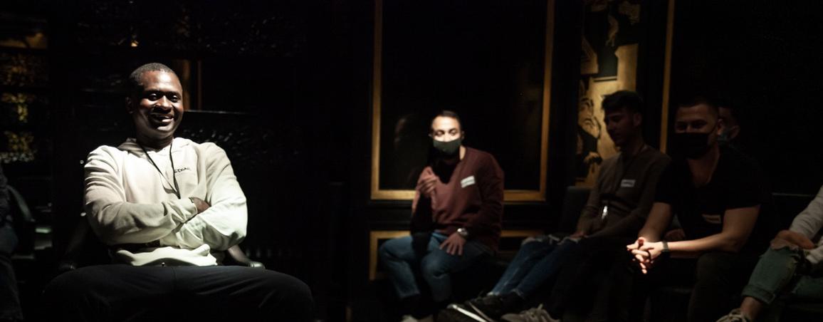 In a dark room a man is on the left lit from above. He is sat down wearing a grey top and smiling. You can see other people sat around in the background.