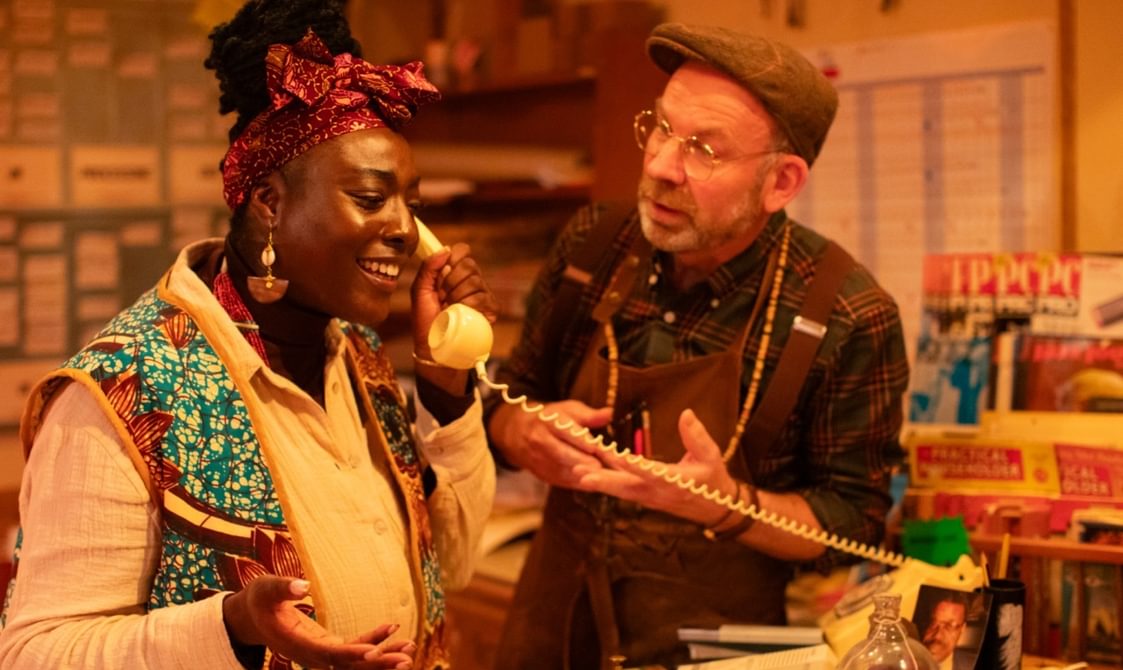 Performers in an immersive theatre show. Enitan is answering the phone and Ged is gesturing at her.
