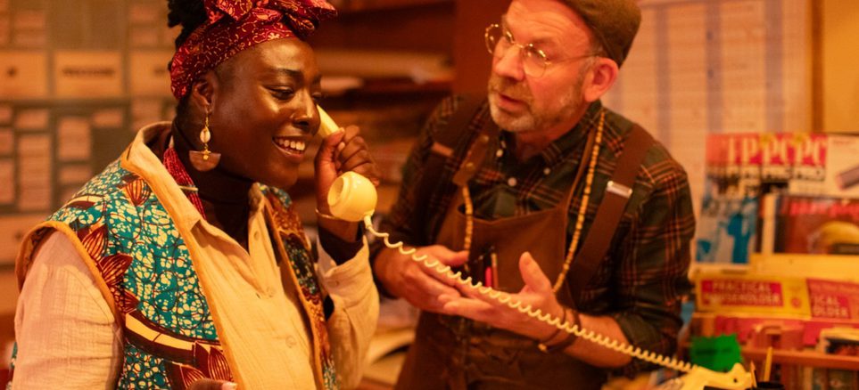 Performers in an immersive theatre show. Enitan is answering the phone and Ged is gesturing at her.