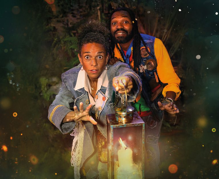 Two people stand in a dark, mystical setting. The person at the front, wearing a denim jacket, reaches towards a glowing lantern, looking surprised. The person behind, in colourful clothes, looks upwards with a cautious expression.