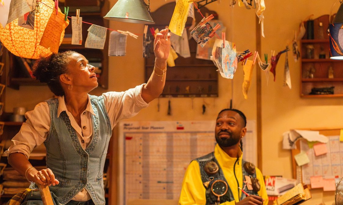 A woman reaches up to pin a note on a string of colourful papers hanging across a cosy, cluttered room, while a man in a yellow zip up jacket and waistcoat looks on, smiling. The walls are decorated with notes and shelves.