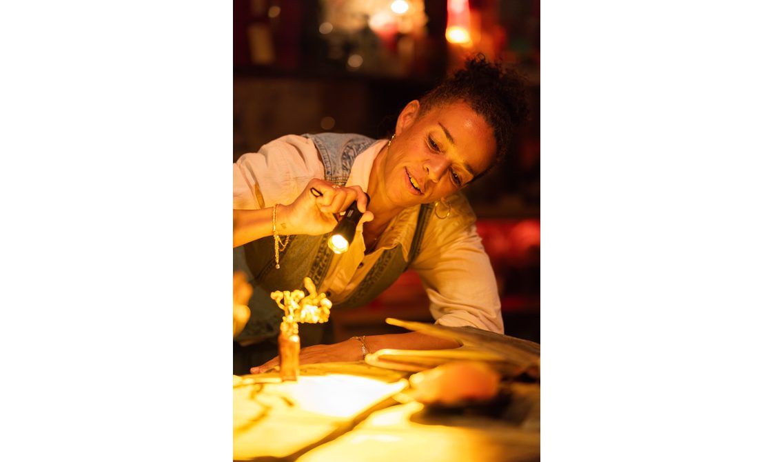 A woman leans over a table, shining a torch on an object. She is focused, wearing a light shirt and denim waistcoat, with warm yellow lighting creating a cosy, intimate atmosphere.