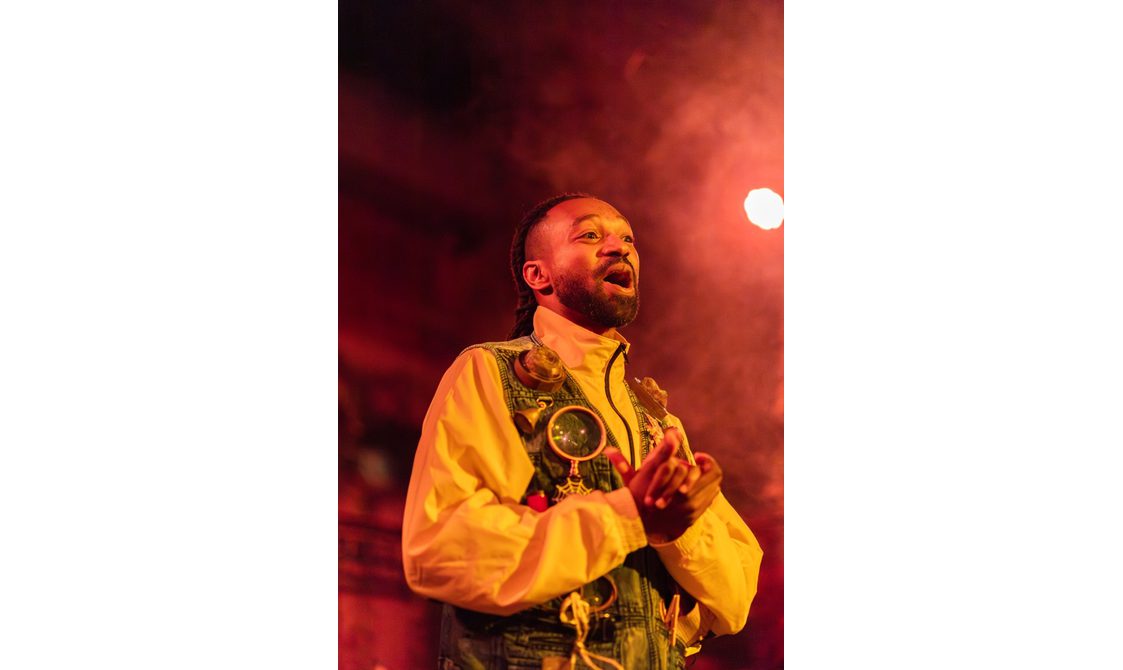 A person wearing a decorated waistcoat and yellow zip up jacket performs passionately under dramatic, warm stage lighting, with hands clasped and mouth open as if speaking.