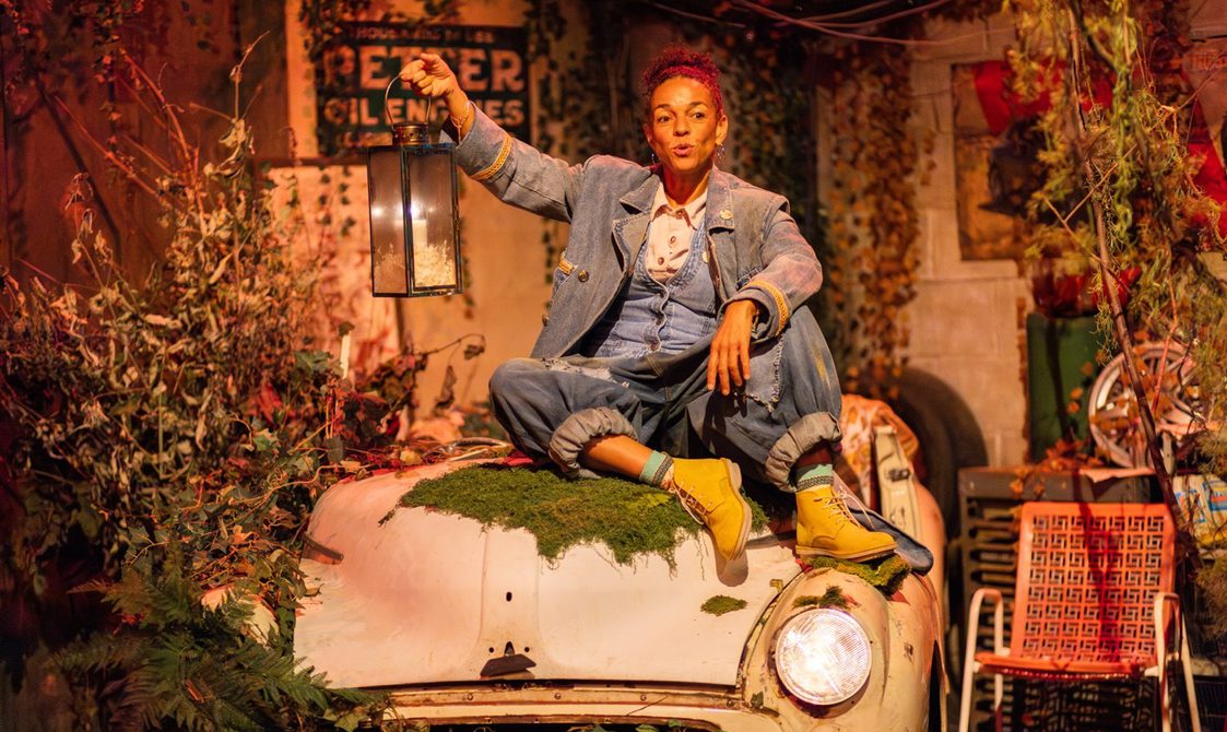 A woman wearing a denim jacket, jeans, and yellow boots, sits on the mossy bonnet of an old car, holding a lantern. The setting is surrounded by plants and vintage signs, creating a rustic atmosphere.