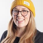 A headshot portrait photograph of a white woman with long blond hair wearing round glasses and a yellow woollen hat.