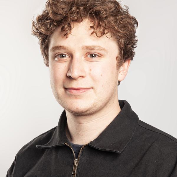Headshot portrait photograph of a white man with short brown curly hair wearing a black zip up jacket.