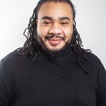 Headshot photograph of a man with shoulder length dreadlocks and a beard. He is wearing a black jumper.