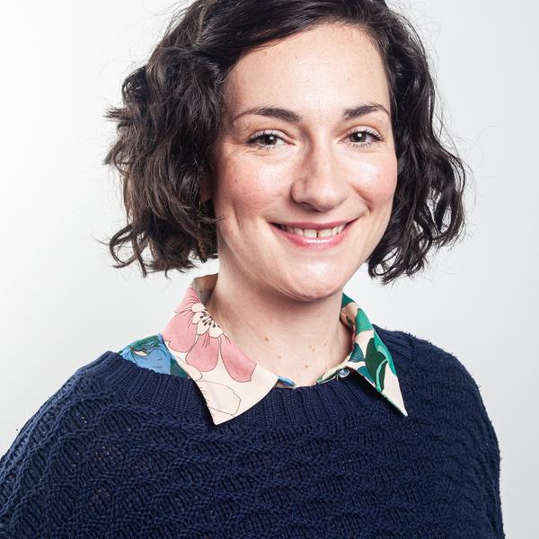 Headshot portrait photo of a white woman with short dark brown curly hair. She is wearing navy blue knitted jumper over a multi-coloured shirt.