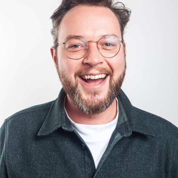 Headshot portrait photo of a white man with short brown hair and a brown and grey beard. He is wearing round glasses and a green shirt over a white t shirt.