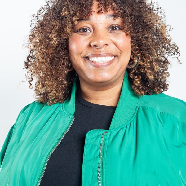 Headshot portrait photo of a woman with curly brown hair. She is wearing a green bomber jacket over a black t shirt.