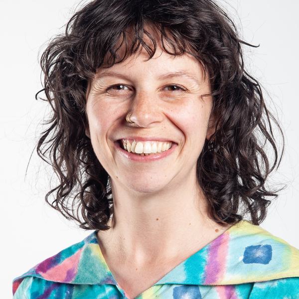 Headshot portrait photo of a white woman with dark brown shoulder length wavy hair and two nose rings. Wearing a multi-coloured shirt.