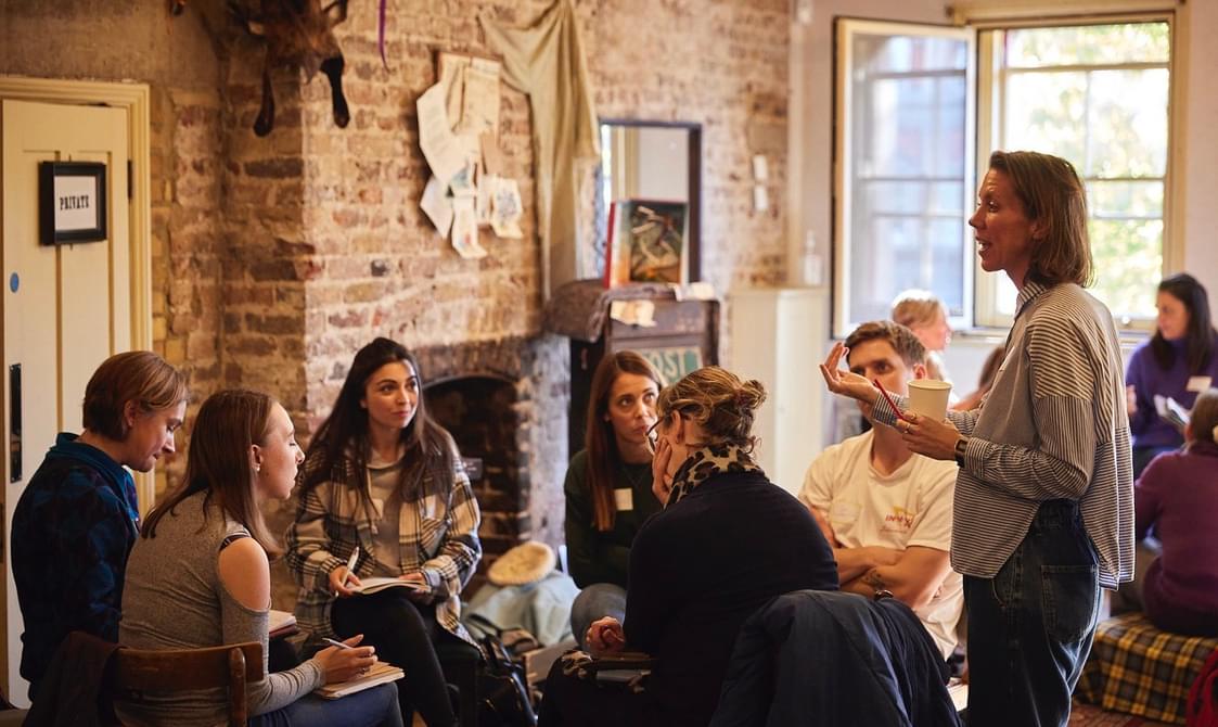 A group of teachers sit in a circle with notebooks and pens. Behind them, is an exposed brick wall and a fireplace.