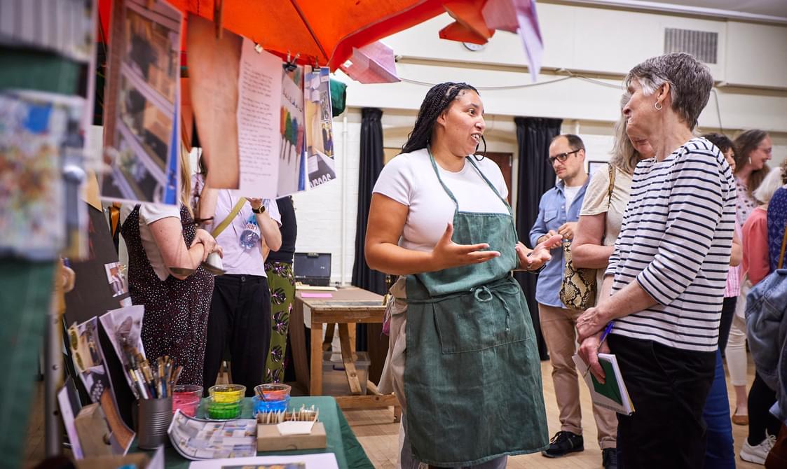 A group of people talking to each other at an indoor marketplace. There are images from immersive learning projects hung up with pegs on a laundry line.