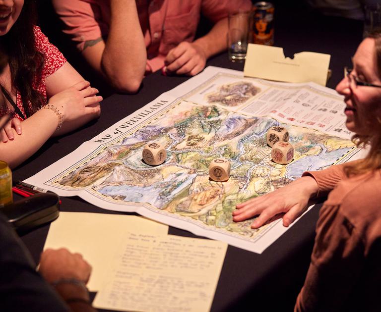A group of people seated around a map of a fictional world . There are some dice on top of it, and other people are writing notes.
