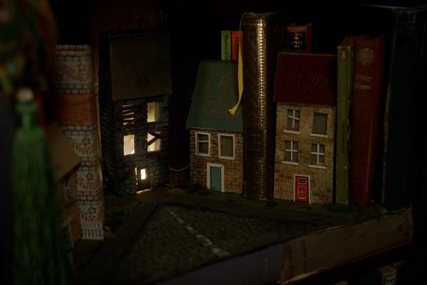 A photograph of some small models of houses on a shelf alongside old leather books. One house has windows that are lit from the inside. The photograph is low light.