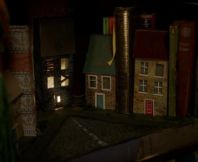 A photograph of some small models of houses on a shelf alongside old leather books. One house has windows that are lit from the inside. The photograph is low light.